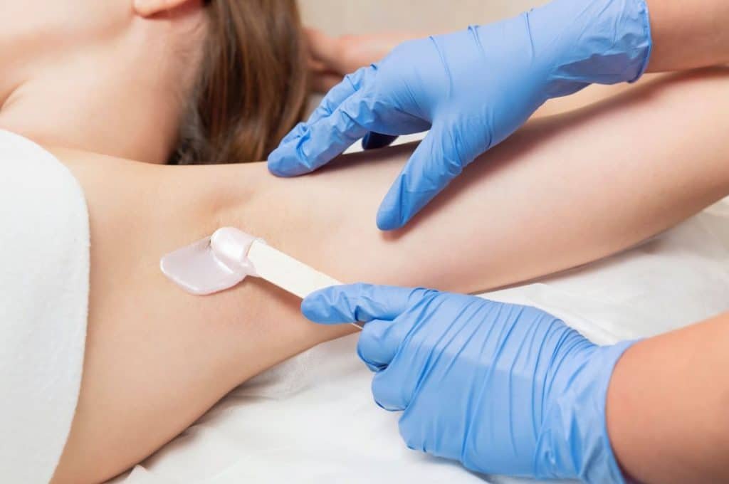 Người đeo găng tay xanh đang thoa sáp tẩy lông lên vùng nách của một người nằm trên giường.