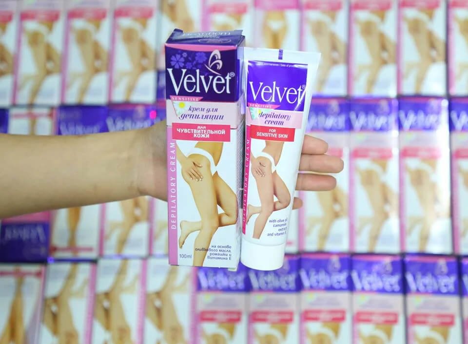 Một bàn tay cầm hộp và tuýp kem tẩy lông Velvet cho da nhạy cảm, nền là nhiều hộp cùng loại.