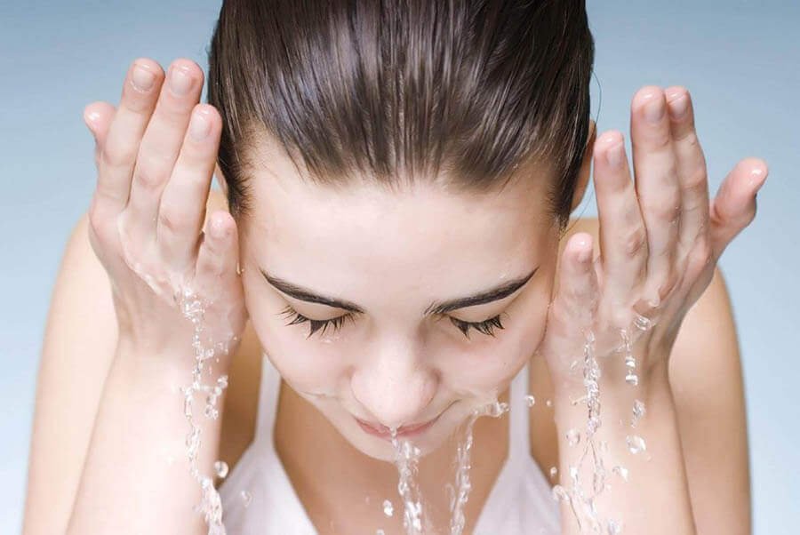 Người đang rửa mặt với nước, tay đưa lên má, mắt nhắm, nền màu xanh nhạt.