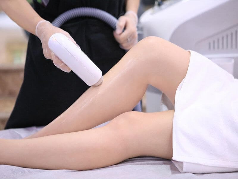 Người đang thực hiện liệu pháp laser trên chân của khách hàng trong một spa, với dụng cụ cầm tay và găng tay bảo vệ.
