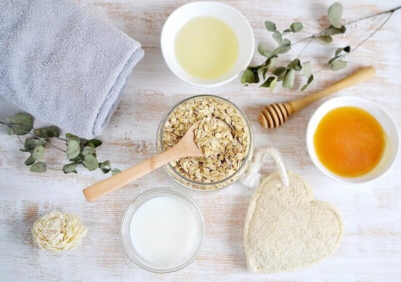Các nguyên liệu thiên nhiên như yến mạch, mật ong, sữa và dầu, với khăn và cành lá trang trí trên bàn.
