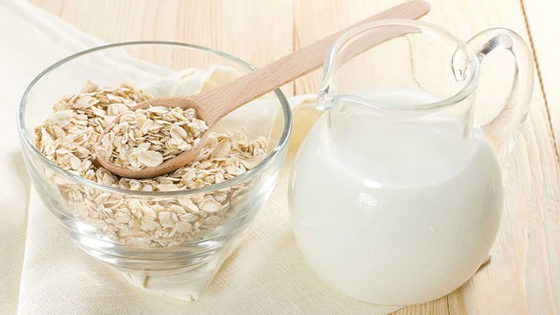 Bát yến mạch và bình sữa trên bàn gỗ sáng màu, có muỗng gỗ trong bát.