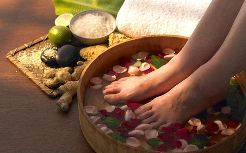 Đôi chân ngâm trong thau gỗ với cánh hoa, bên cạnh là gừng, chanh, muối và khăn trắng.