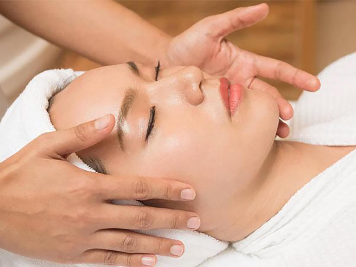 Người đang nằm, được massage mặt, quấn khăn trắng quanh đầu trong không gian thư giãn.