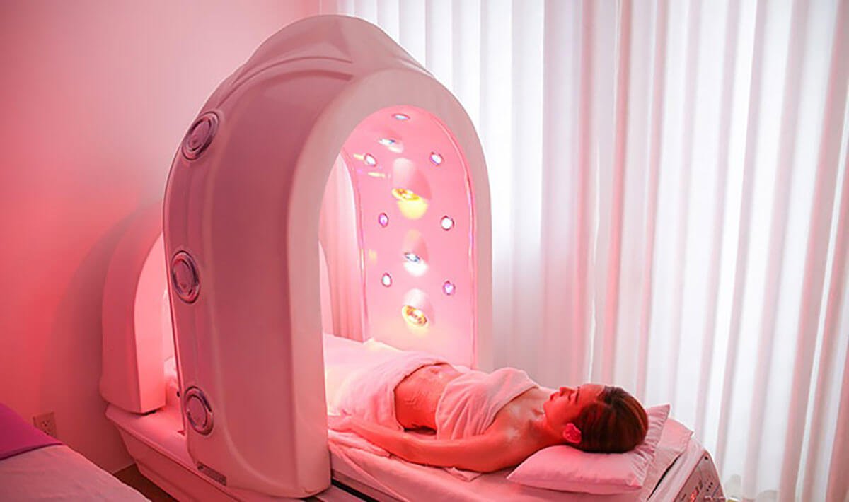 Người nằm thư giãn trong máy chăm sóc sức khỏe với ánh sáng màu hồng và trắng, bên trong phòng có rèm trắng.