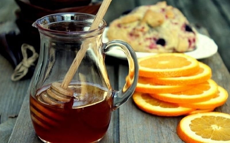 Bình mật ong với que khuấy, lát cam và bánh trên bàn gỗ.
