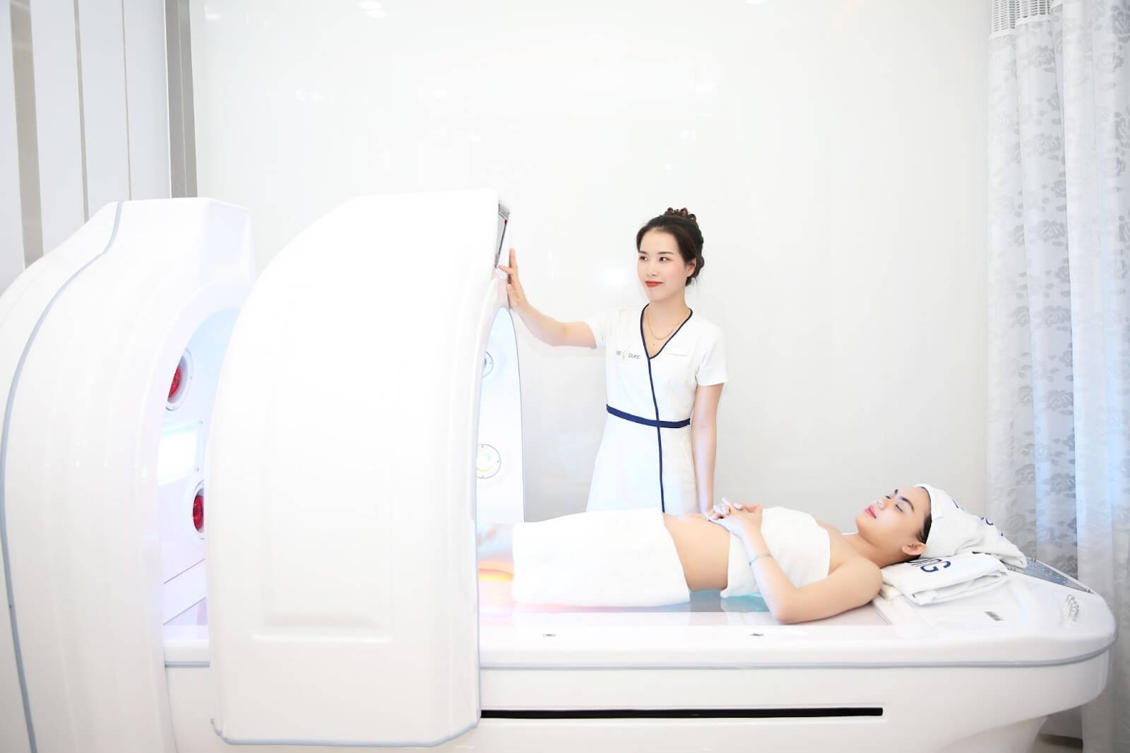 Nhân viên thẩm mỹ viện đứng cạnh máy chăm sóc da, bên cạnh là khách hàng nằm thư giãn trên giường trị liệu.