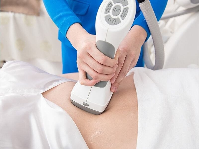 Người cầm thiết bị massage bụng trên cơ thể nằm, mặc áo trắng, trong môi trường chăm sóc sức khỏe.