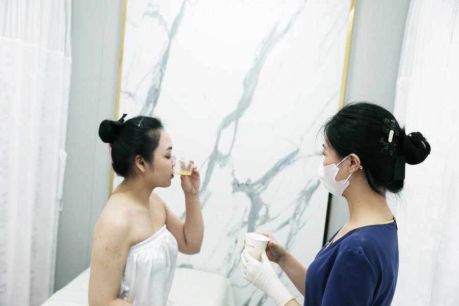 Hai người đang trao đổi cốc nước trong phòng có tường đá cẩm thạch, một người mặc đồng phục và đeo khẩu trang.