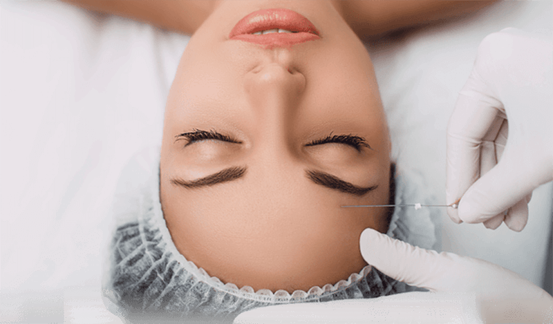 Người đeo găng tay chuẩn bị tiêm lên trán của một người đang nằm, đội mũ y tế.