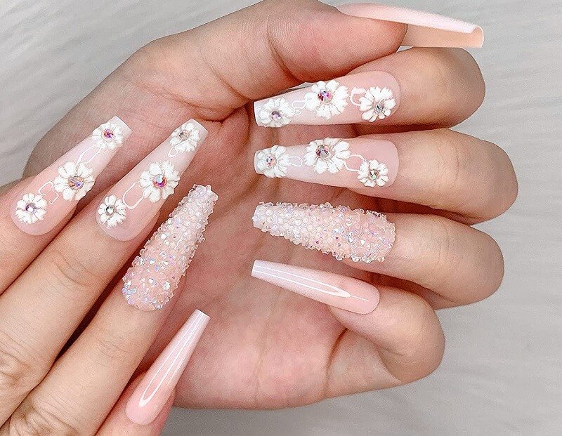 Bàn tay với móng tay dài, trang trí hoa và hạt lấp lánh trên nền màu hồng nhạt.