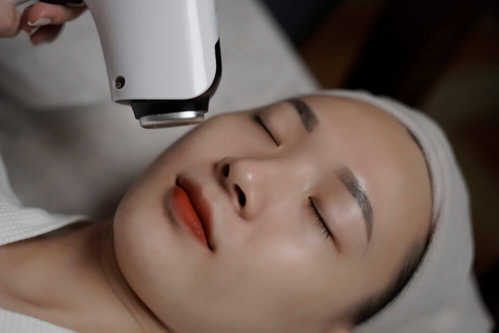 Người đang nằm nhắm mắt, nhận liệu trình chăm sóc da mặt bằng thiết bị công nghệ cao.