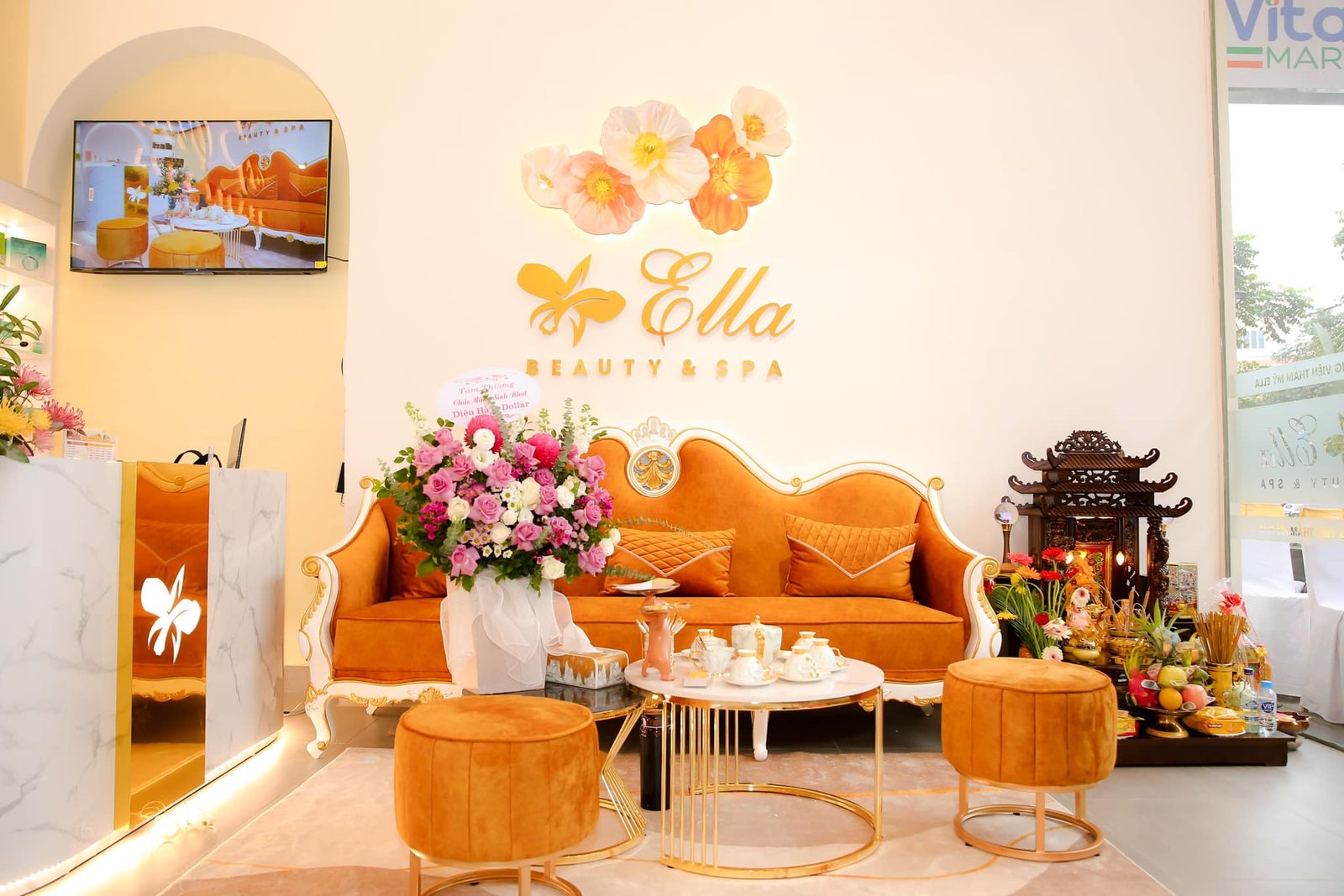 Top 10+ Tiệm Nail Quận 11 Đẹp, Chuyên Nghiệp Được Yêu Thích Hiện Nay 5 Phòng tiếp khách spa với ghế sofa màu cam, bàn trà, hoa tươi và kệ thờ trang trí.