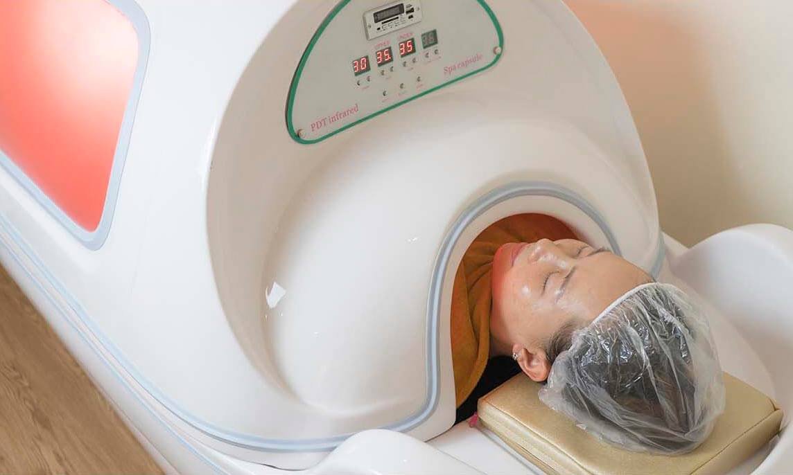 Người nằm thư giãn trong máy xông hơi toàn thân, đầu ngẩng ra ngoài, tóc được bọc trong mũ nhựa.