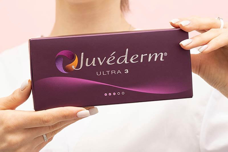 Người cầm hộp sản phẩm màu tím có chữ "Juvéderm Ultra 3" trên nền hồng.