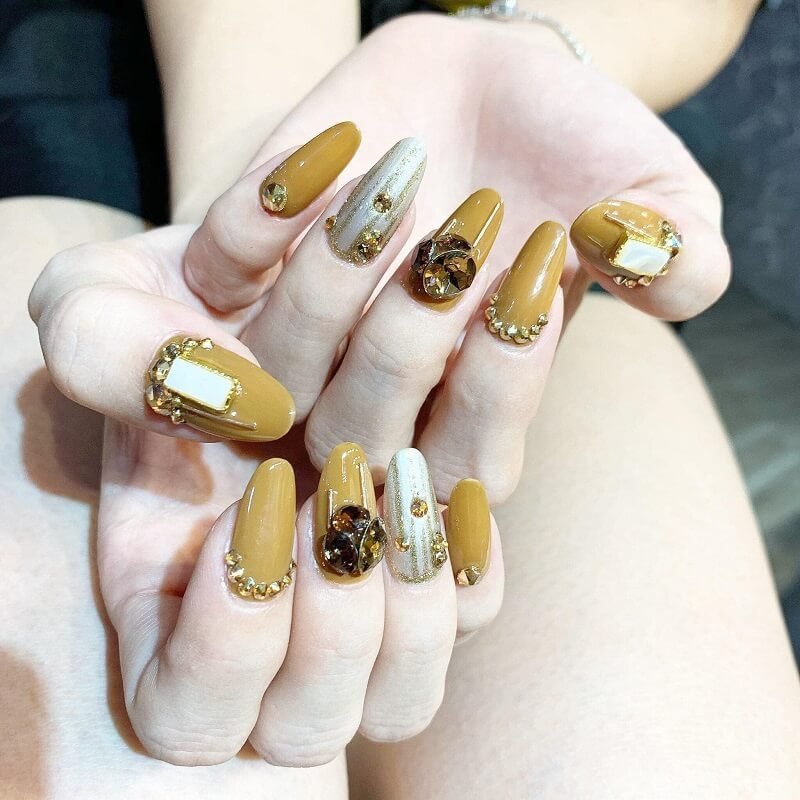 Bàn tay với móng tay sơn màu nâu và trang trí đá quý lấp lánh, tạo kiểu nghệ thuật độc đáo và tinh tế.