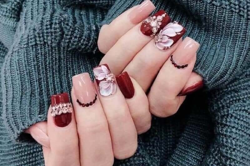 Bàn tay với móng tay được sơn màu đỏ và hồng, trang trí hoa và đính đá, trên nền áo len màu xanh.