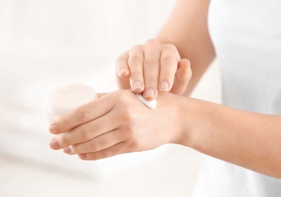 Người đang thoa kem từ một hũ nhỏ lên cổ tay, nền màu sáng.