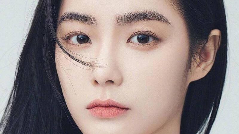 Chân dung một người với mái tóc đen dài, ánh mắt nhìn thẳng, và đôi môi nhẹ nhàng, trên nền màu sáng.