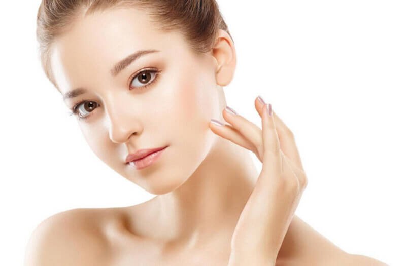 Người phụ nữ với làn da mịn màng, tóc búi cao, đang chạm nhẹ vào má trên nền trắng.