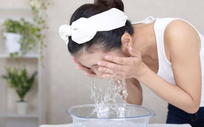 Người phụ nữ rửa mặt bằng nước từ chậu, đeo băng đô trắng, đứng trong phòng có cây xanh.