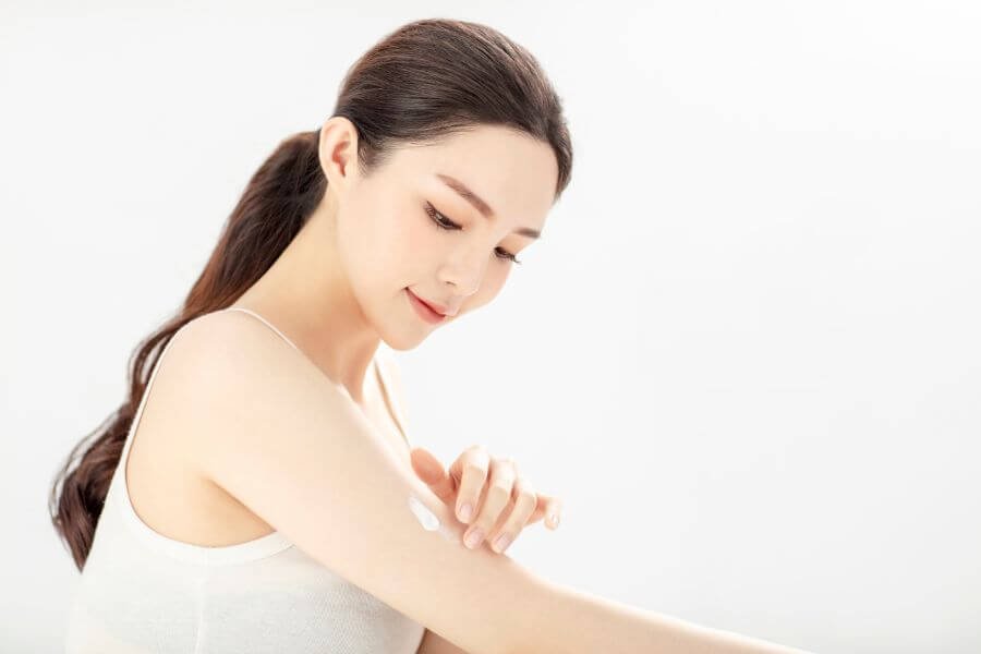 Người phụ nữ mặc áo trắng đang bôi kem dưỡng da lên cánh tay, nền trắng đơn giản.