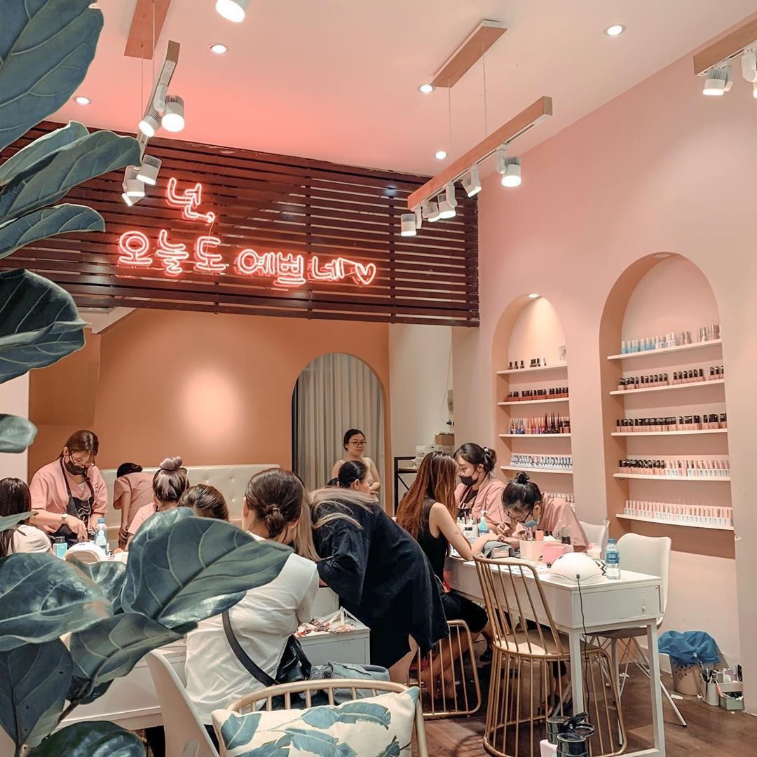 Top 10+ Tiệm Nail Quận 11 Đẹp, Chuyên Nghiệp Được Yêu Thích Hiện Nay 3 Một tiệm nail với khách hàng và nhân viên đang làm móng, không gian nội thất màu hồng, có đèn neon trên tường.