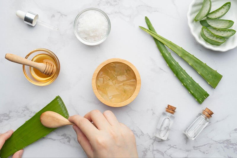 Người đang dùng muỗng gỗ lấy gel từ lá nha đam trên nền đá cẩm thạch, xung quanh có mật ong và nguyên liệu tự nhiên.