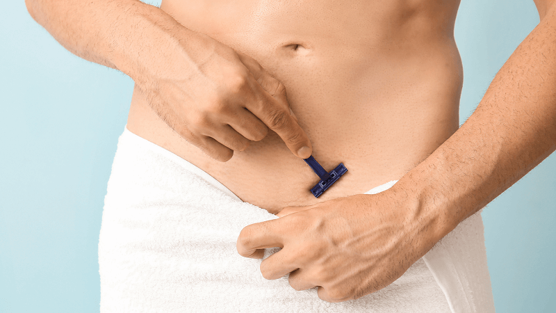 Người cầm dao cạo màu xanh chuẩn bị cạo lông vùng bụng, quấn khăn trắng quanh eo, nền xanh nhạt.