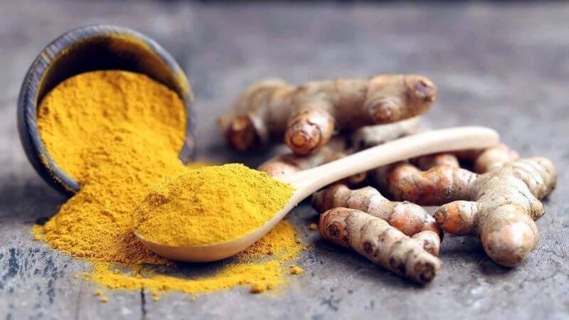 Bột nghệ vàng trong muỗng gỗ và củ nghệ tươi trên nền gỗ xám.