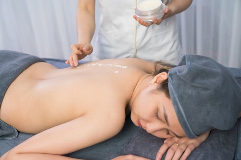 Người nằm thư giãn trong spa, có khăn che đầu, người khác thoa kem lên lưng, tạo cảm giác thư giãn.