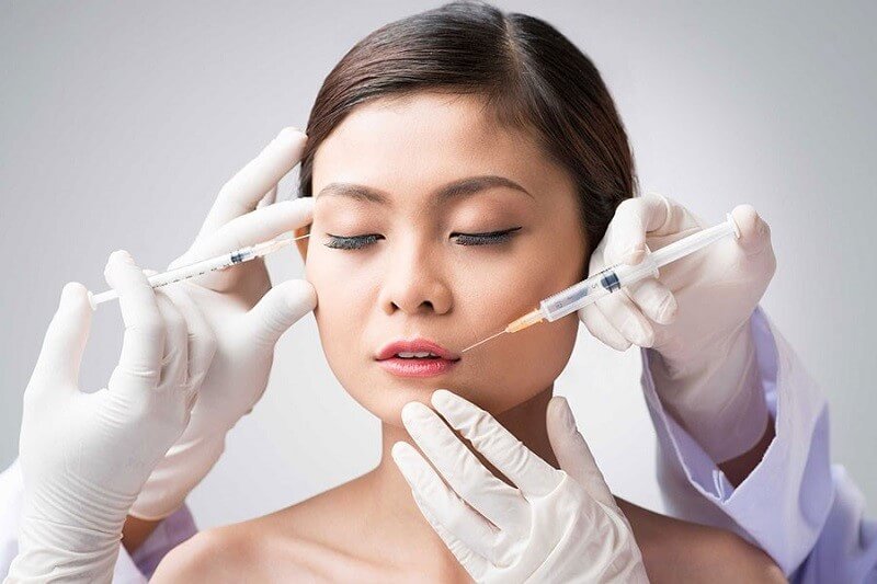Người được tiêm chất làm đầy vào mặt bởi hai tay đeo găng, mắt nhắm, trong bối cảnh trắng.