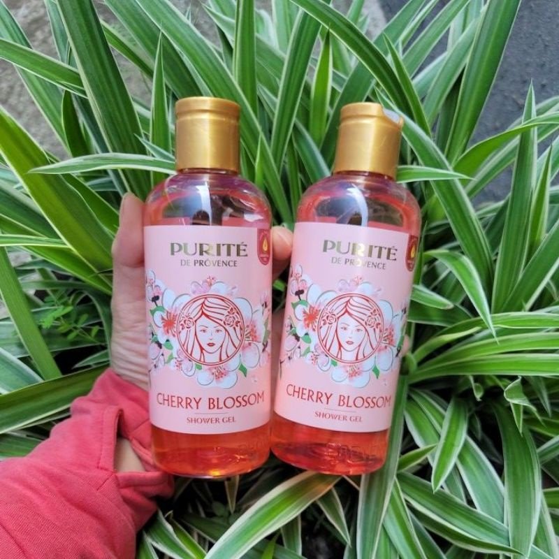 Hai chai sữa tắm Purité de Provence hương hoa anh đào, nắp màu vàng, được cầm trên nền lá cây xanh.