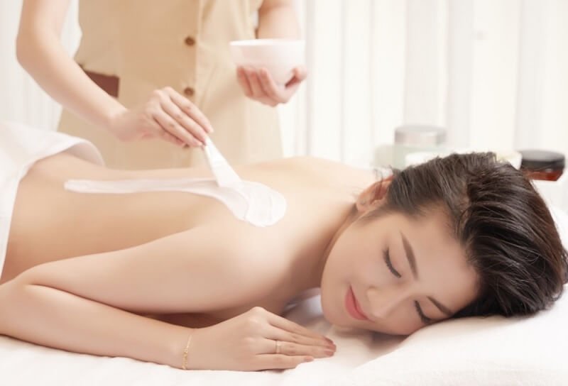 Người phụ nữ nằm thư giãn trên giường spa, đang được thoa kem dưỡng lên lưng bởi một nhân viên.