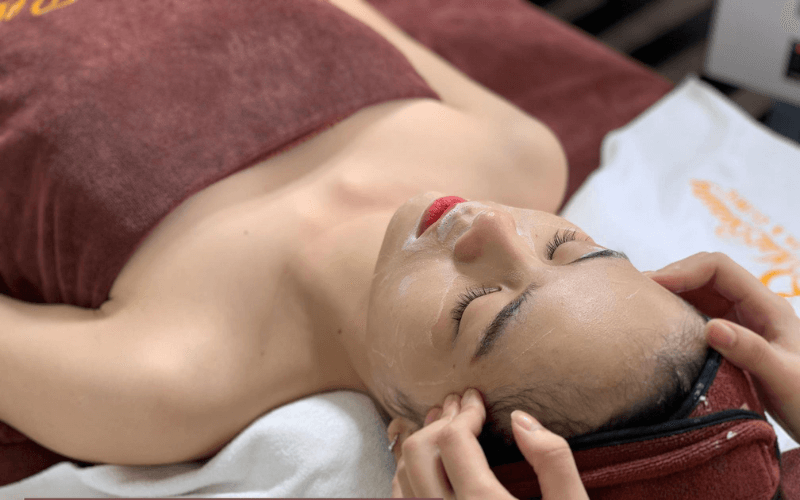 Người đang được massage mặt, nằm trên giường spa, khăn che người, không gian thư giãn.