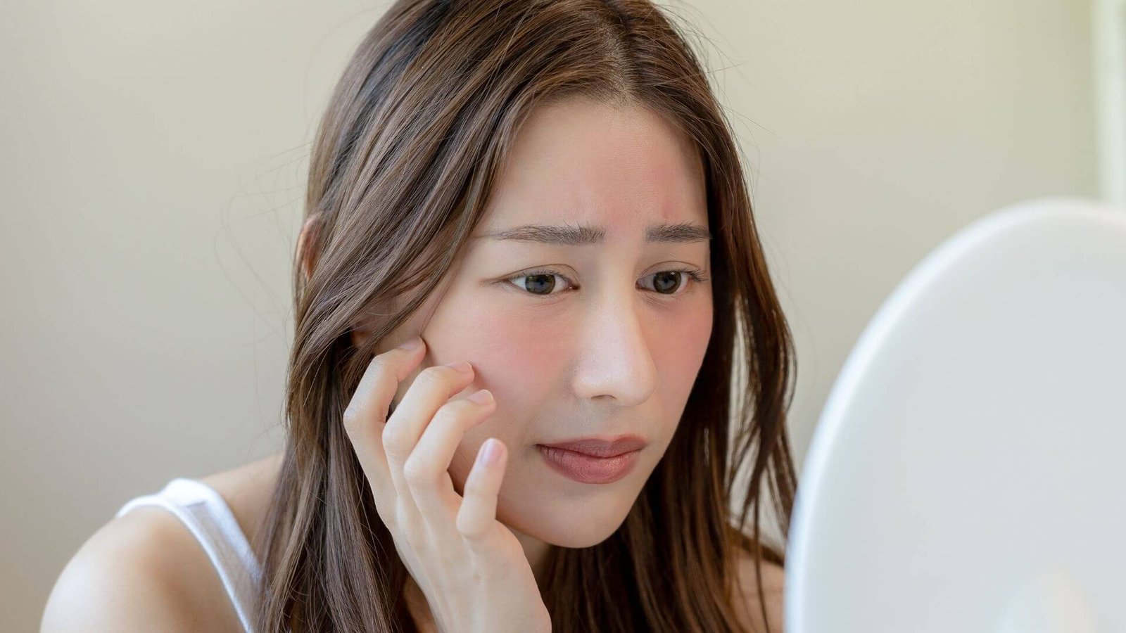 Người phụ nữ nhìn vào gương, chạm vào má, biểu cảm lo lắng, có dấu hiệu đỏ trên da mặt.
