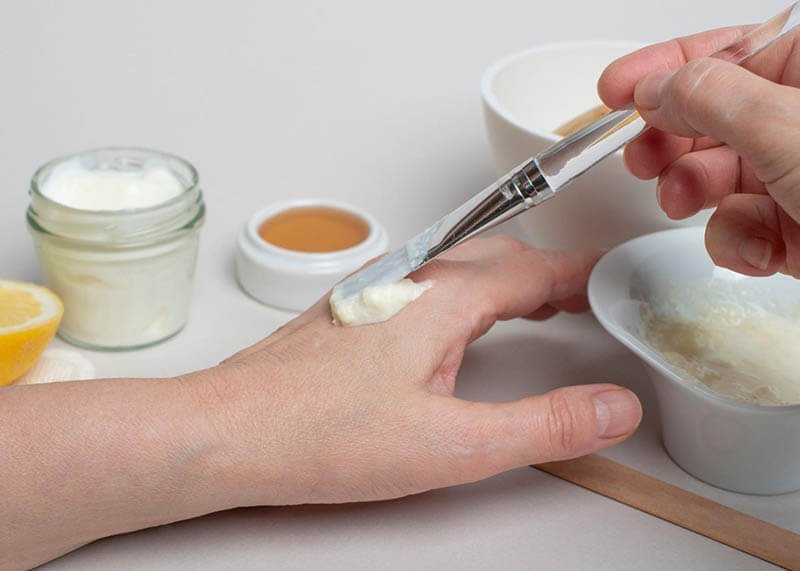 Một bàn tay đang được bôi kem bằng cọ; xung quanh có các lọ chứa kem và chanh.