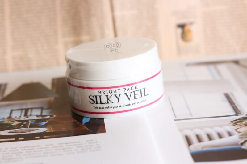 Hũ kem dưỡng da màu trắng, nhãn hiệu "Silky Veil", đặt trên nền sách báo mở.