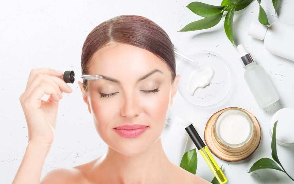 Người phụ nữ dùng ống nhỏ giọt thoa serum lên mặt, xung quanh là sản phẩm chăm sóc da và lá cây.