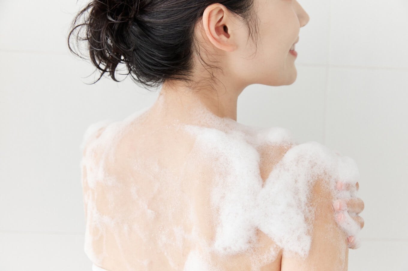 Người đang tắm, lưng phủ đầy bọt xà phòng, tóc buộc gọn, trên nền tường trắng.