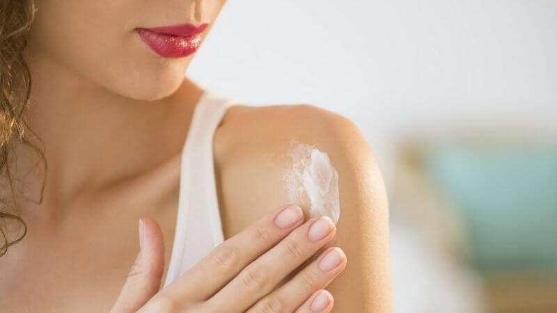 Người phụ nữ thoa kem dưỡng da lên vai, mặc áo trắng, môi đỏ nổi bật.