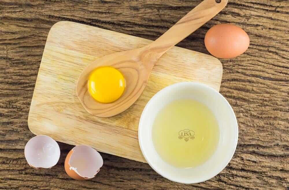 Lòng đỏ trứng trên thìa gỗ, lòng trắng trong bát, vỏ trứng vỡ trên thớt gỗ, bên cạnh trứng nguyên.