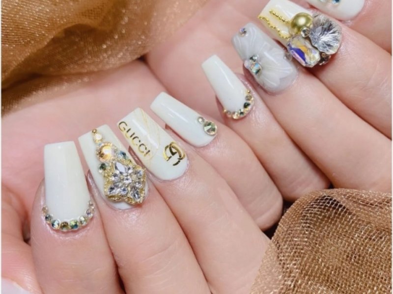 Bàn tay với móng tay sơn trắng, trang trí họa tiết và đính đá lấp lánh, trên nền vải màu nâu sáng.