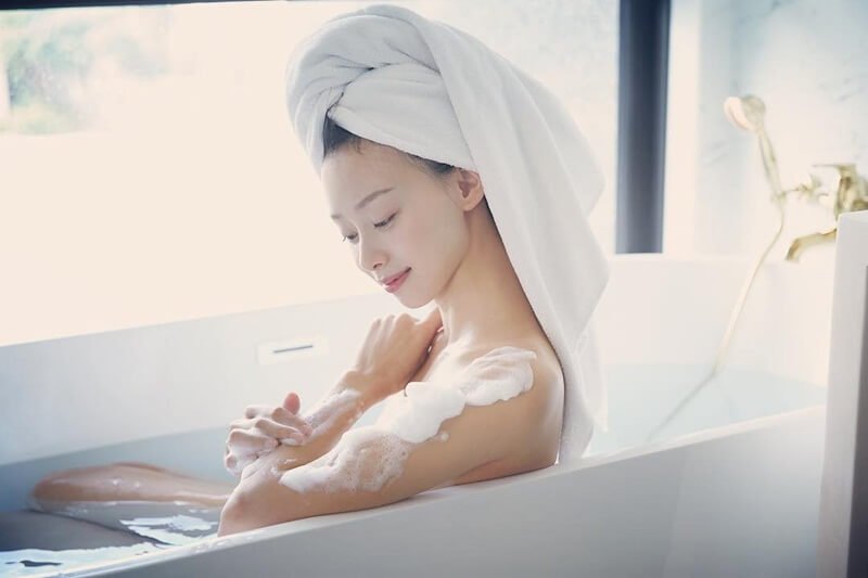 Người phụ nữ quấn khăn tắm ngồi trong bồn tắm, tạo bọt xà phòng trên cánh tay, ánh sáng từ cửa sổ chiếu vào.
