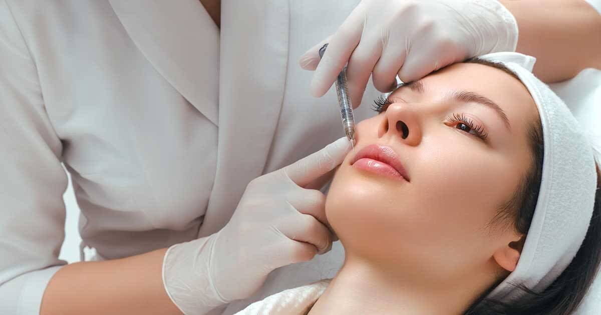 Người đeo găng tay thực hiện tiêm thẩm mỹ lên khuôn mặt người nằm trên giường, đầu đội băng đô trắng.