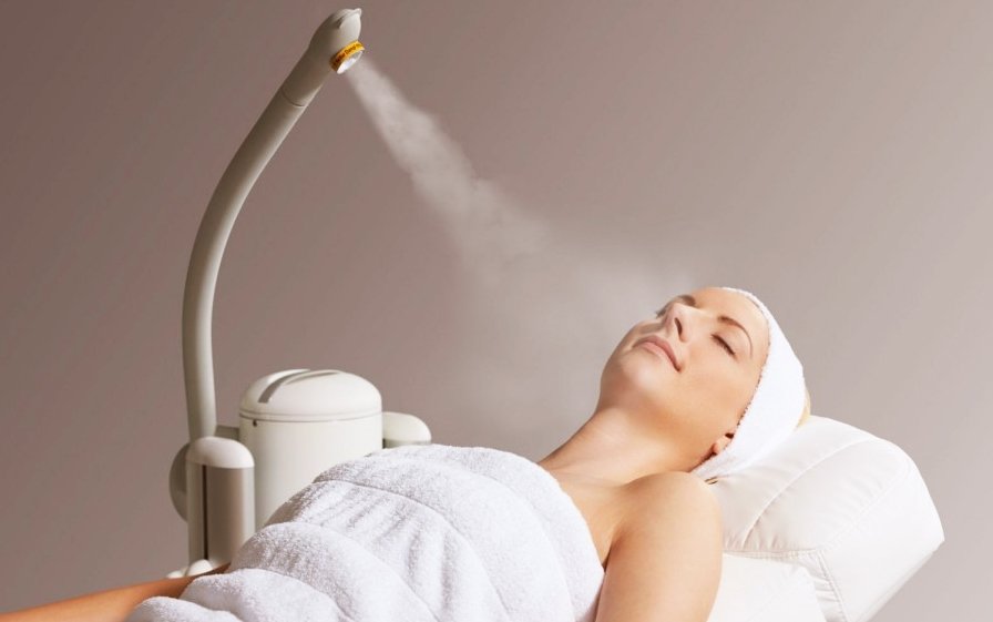 Người nằm trên giường spa, được phủ khăn, máy phun sương đang hoạt động gần khuôn mặt.
