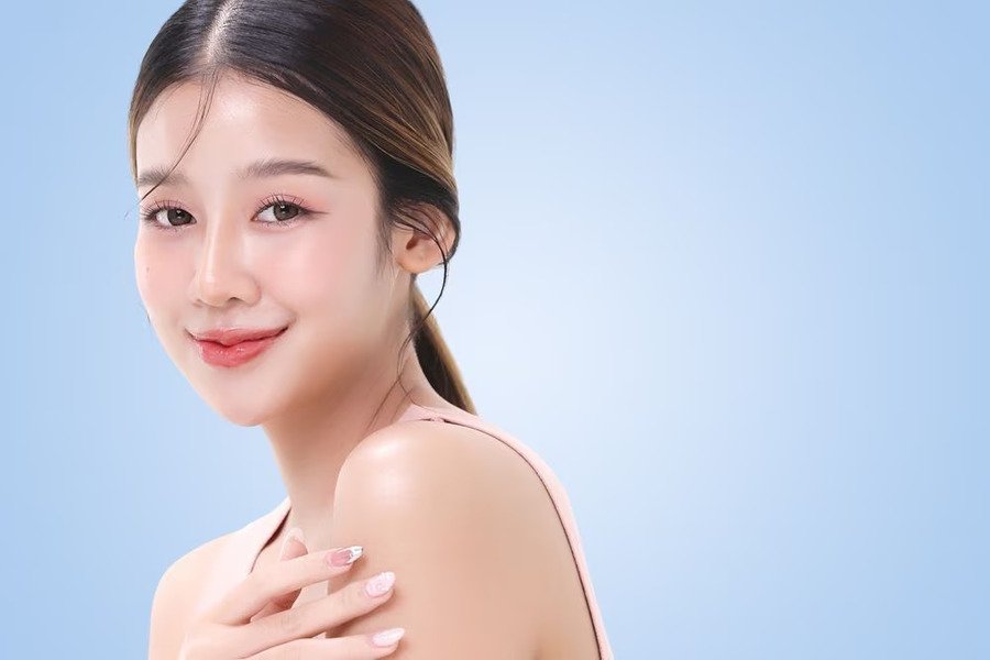Một người đang mỉm cười, tóc buộc gọn, nền xanh nhạt, mặc áo hở vai, tay đặt nhẹ trên vai.